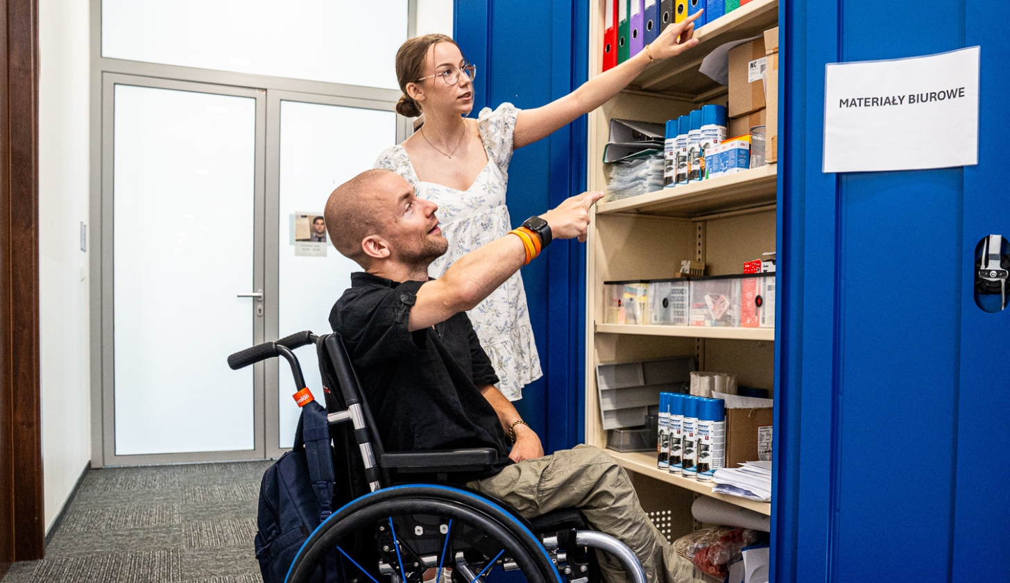 Mężczyzna na wózku wskazuje na przedmioty w szafie biurowej. Obok niego stoi kobieta.