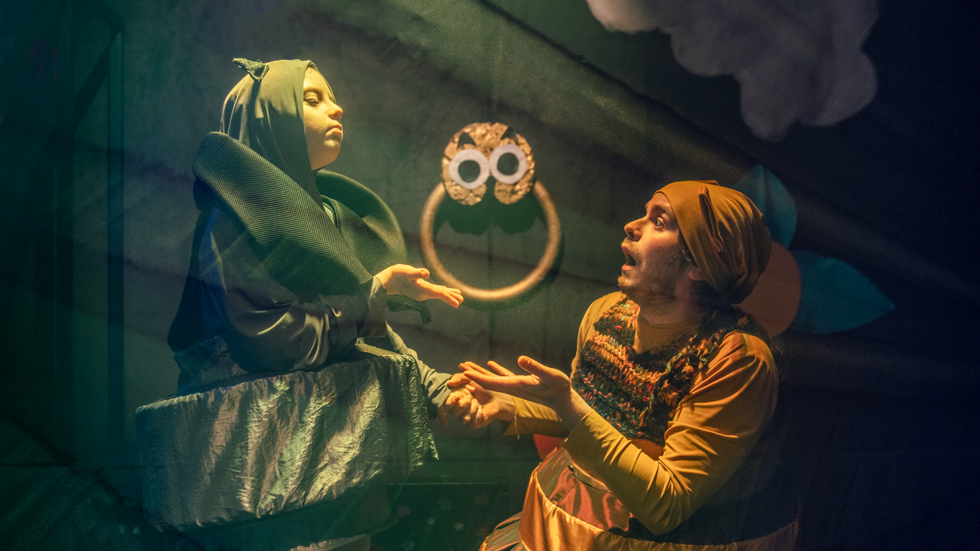 Na scenie dwie postacie, ubrane są w kostiumy. Oświetla je światło. W tle widać element dekoracji.