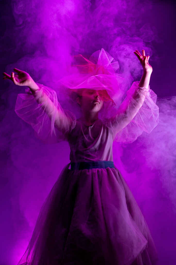 Kobieta ubrana w suknię, unosi w górę ręce. Jej postać owiana jest dymem i oświetlona fioletowym światłem.