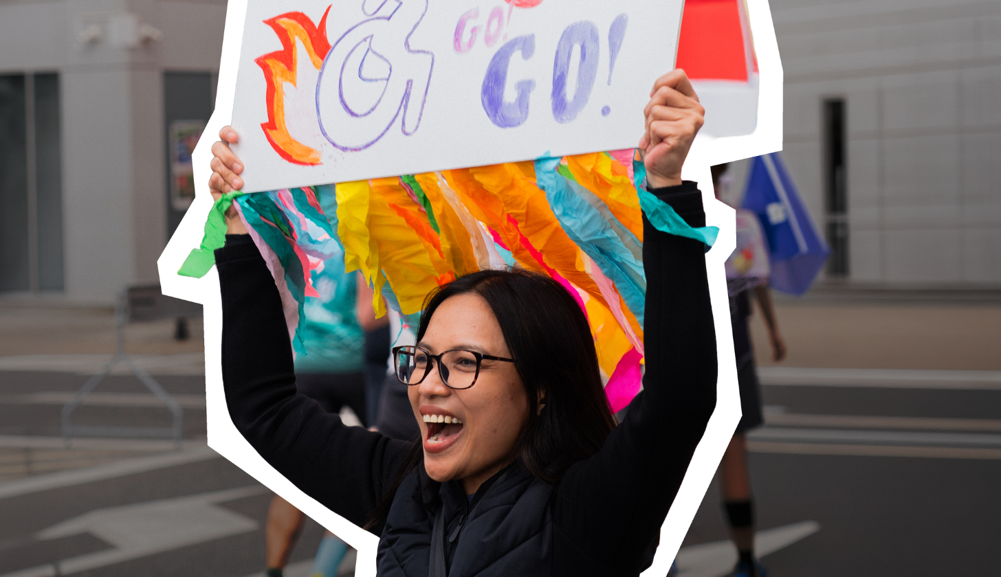 Kobieta w okularach ma otwarte usta i trzyma nad głową transparent.