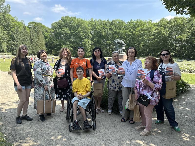 Grupa ludzi stoi w parku na tle zieleni i drzew. Jedna osoba siedzi na wózku inwalidzkim. Kilka osób trzyma w rękach książki oraz torby. Zdjęcie zostało wykonane w słoneczny dzień, w otoczeniu ścieżek i roślinności.
