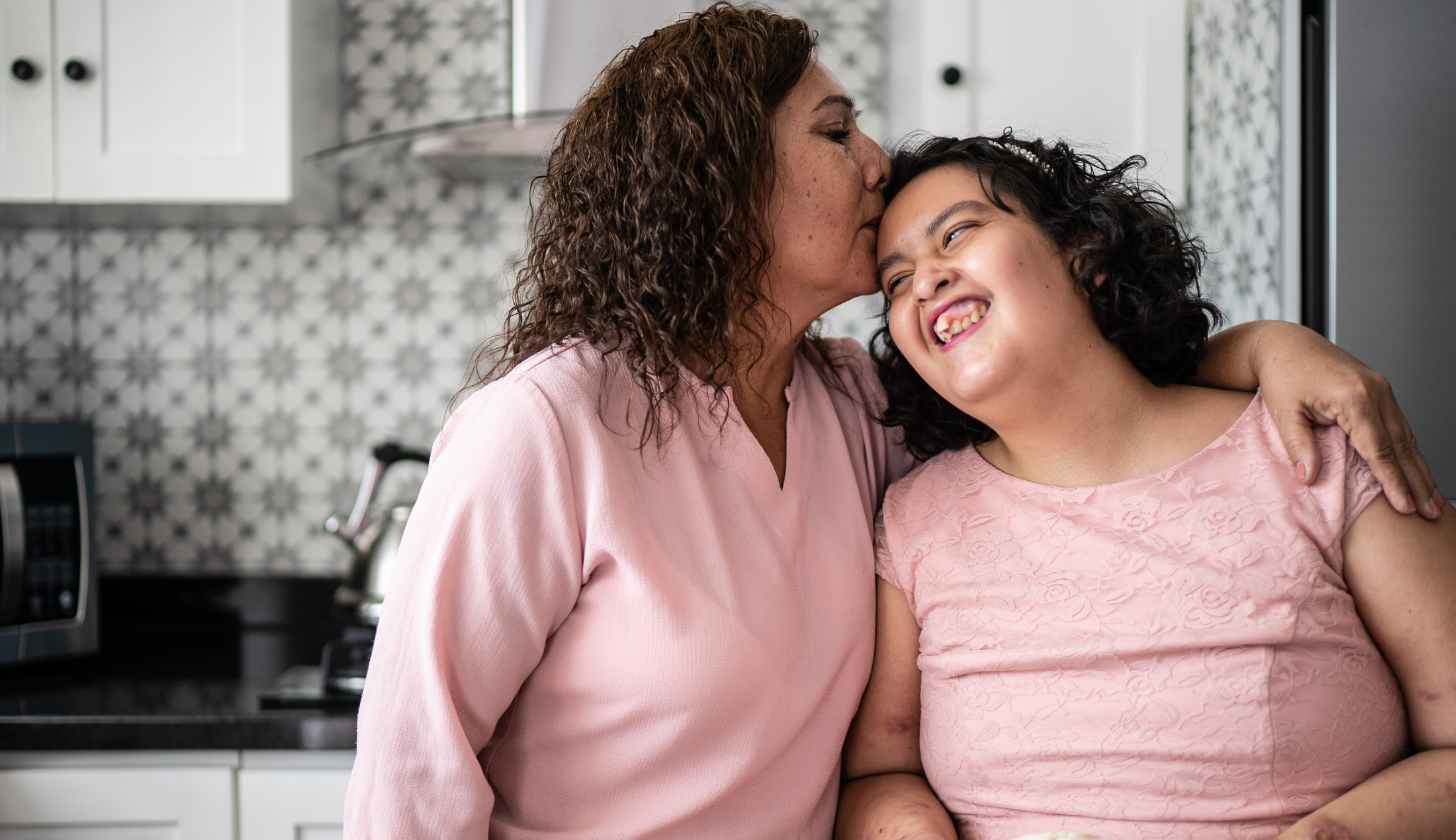 kobieta obejmuje nastoletnią córkę i daje jej buziaka w głowę obie są ubrane na różowo