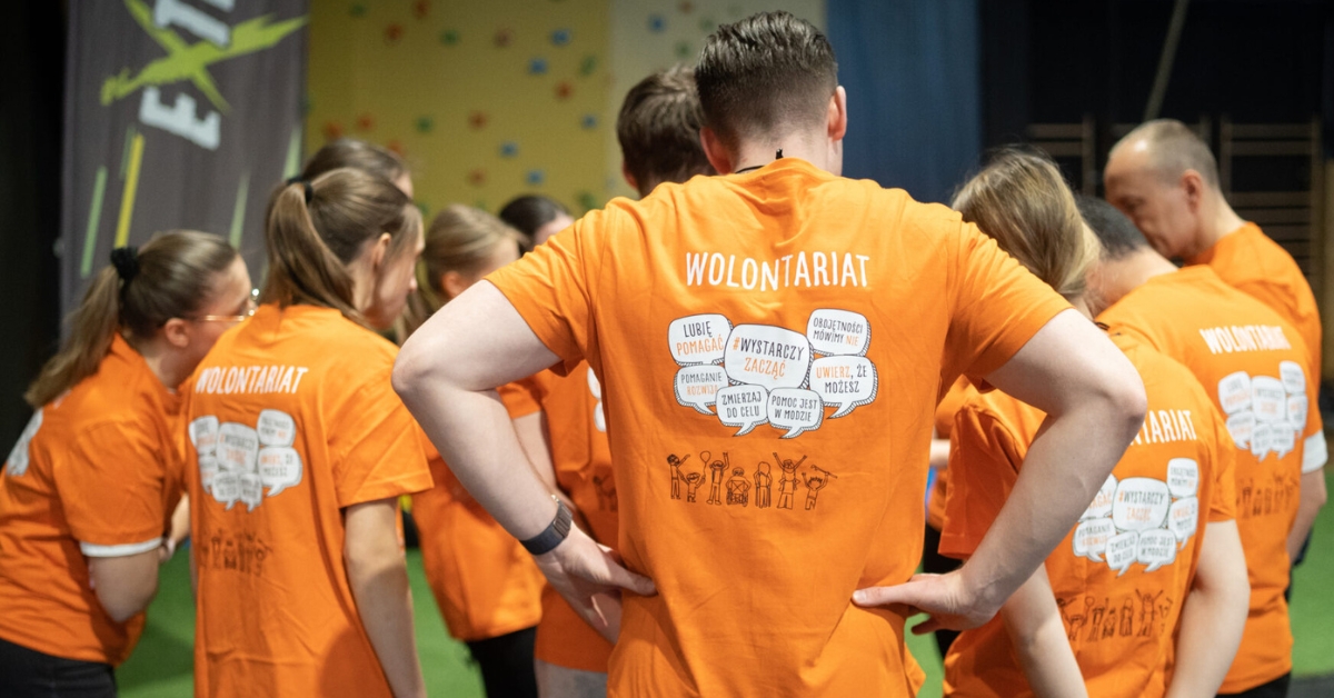 Osoby stojące tyłem ubrane są w pomarańczowe koszulki z napisem wolontariat.