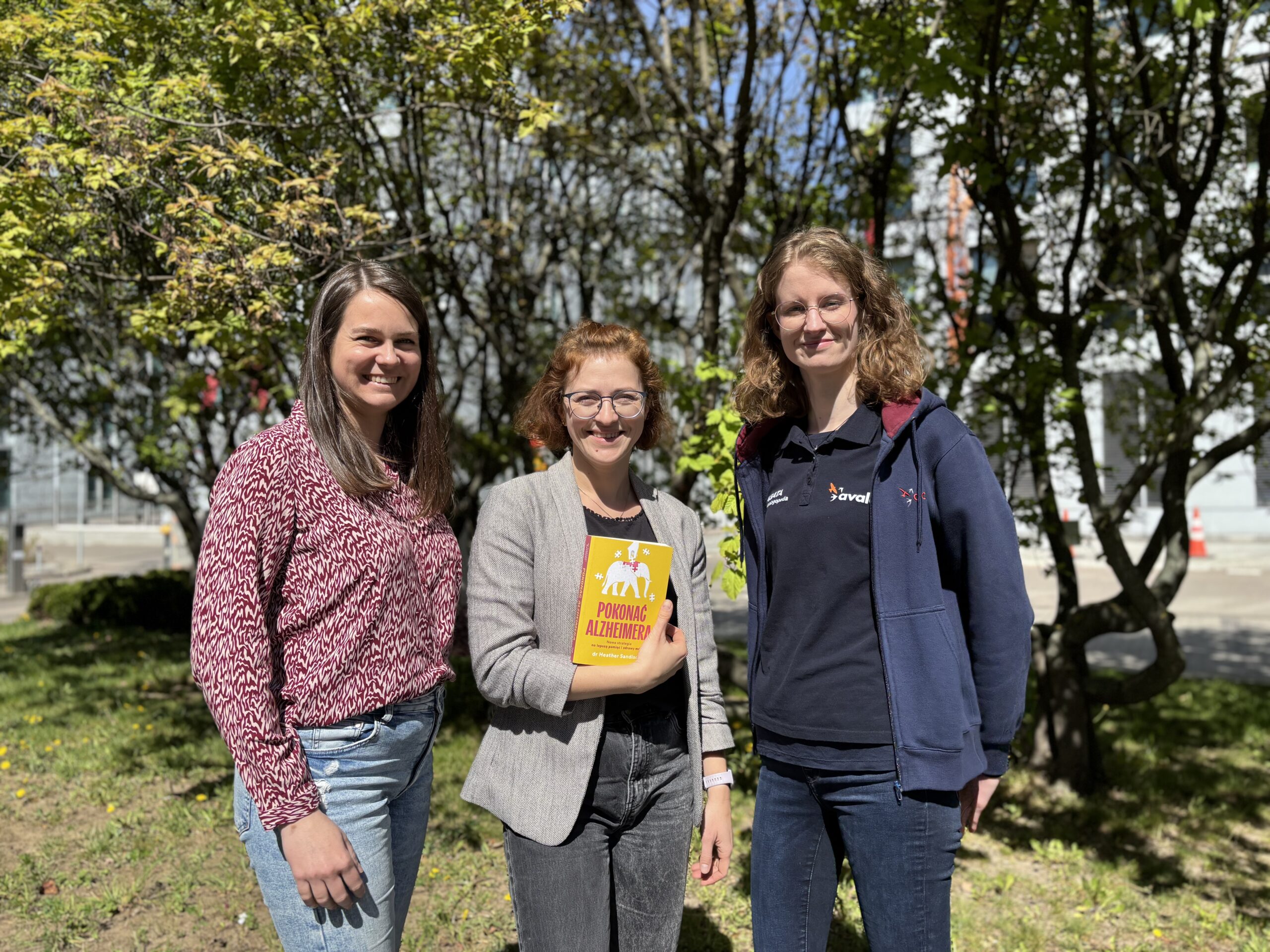 Trzy kobiety stoją obok siebie, jedna z nich trzyma w ręku książkę pokazując okładkę. W tle są drzewa z zielonymi liśćmi.