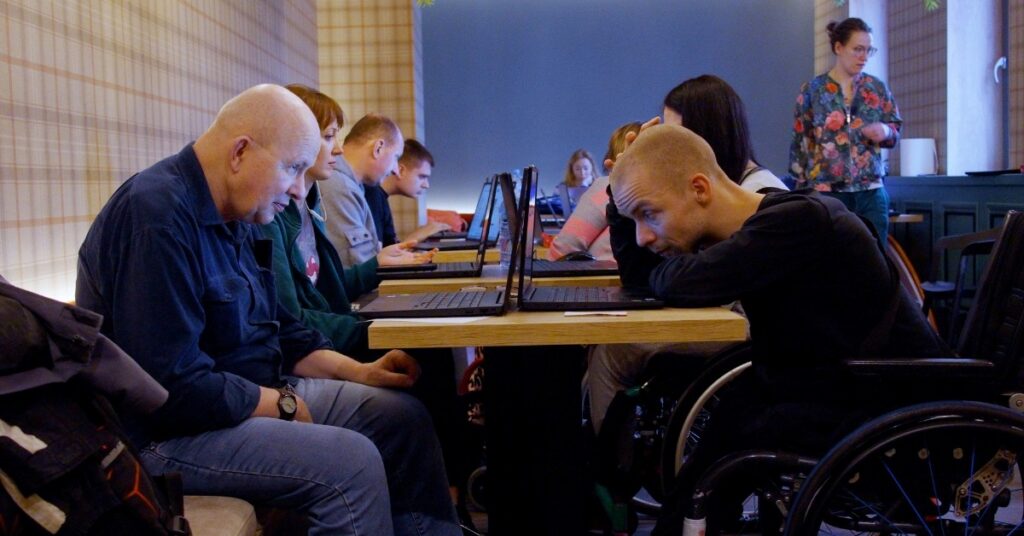 Dwóch mężczyzn siedzi przy stoliku po przeciwnych stronach. Patrząc w ekrany laptopów. W tle inni siedzący ludzie.