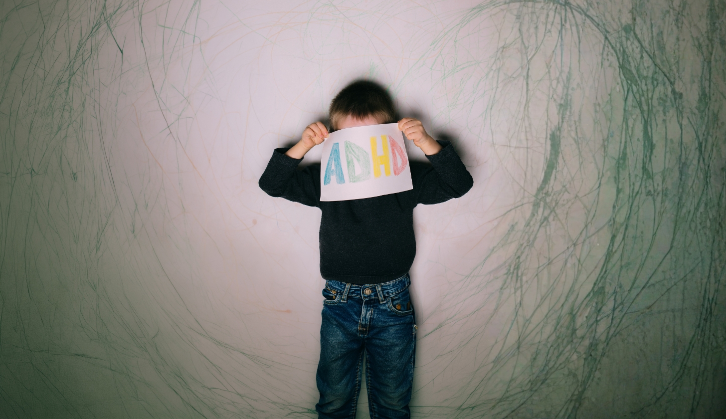 Dziecko stoi przy ścianie i zakrywa swoją twarz kartką z kolorowym napisem ADHD.