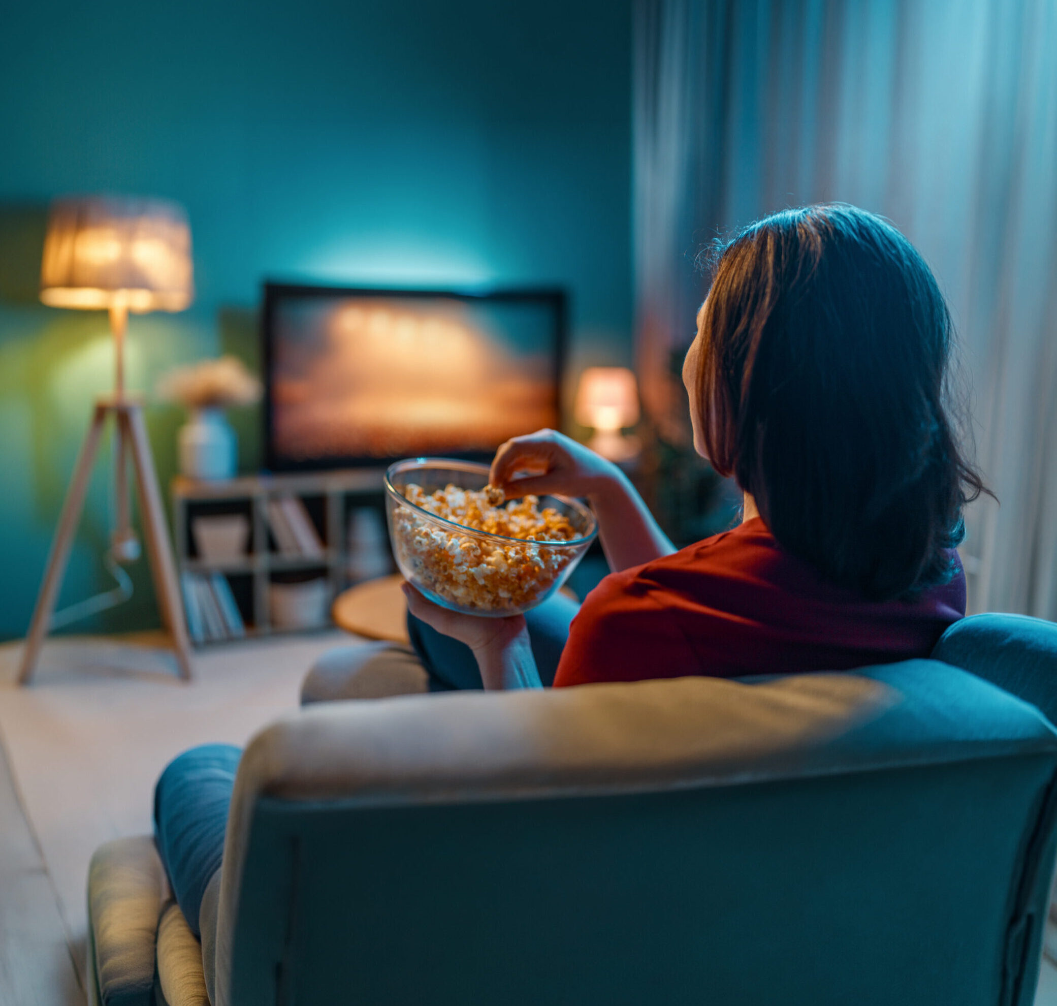 W pokoju kobieta jest odwrócona tyłem, siedzi na fotelu, w misce ma popcorn. W tle widać telewizor i lampkę.