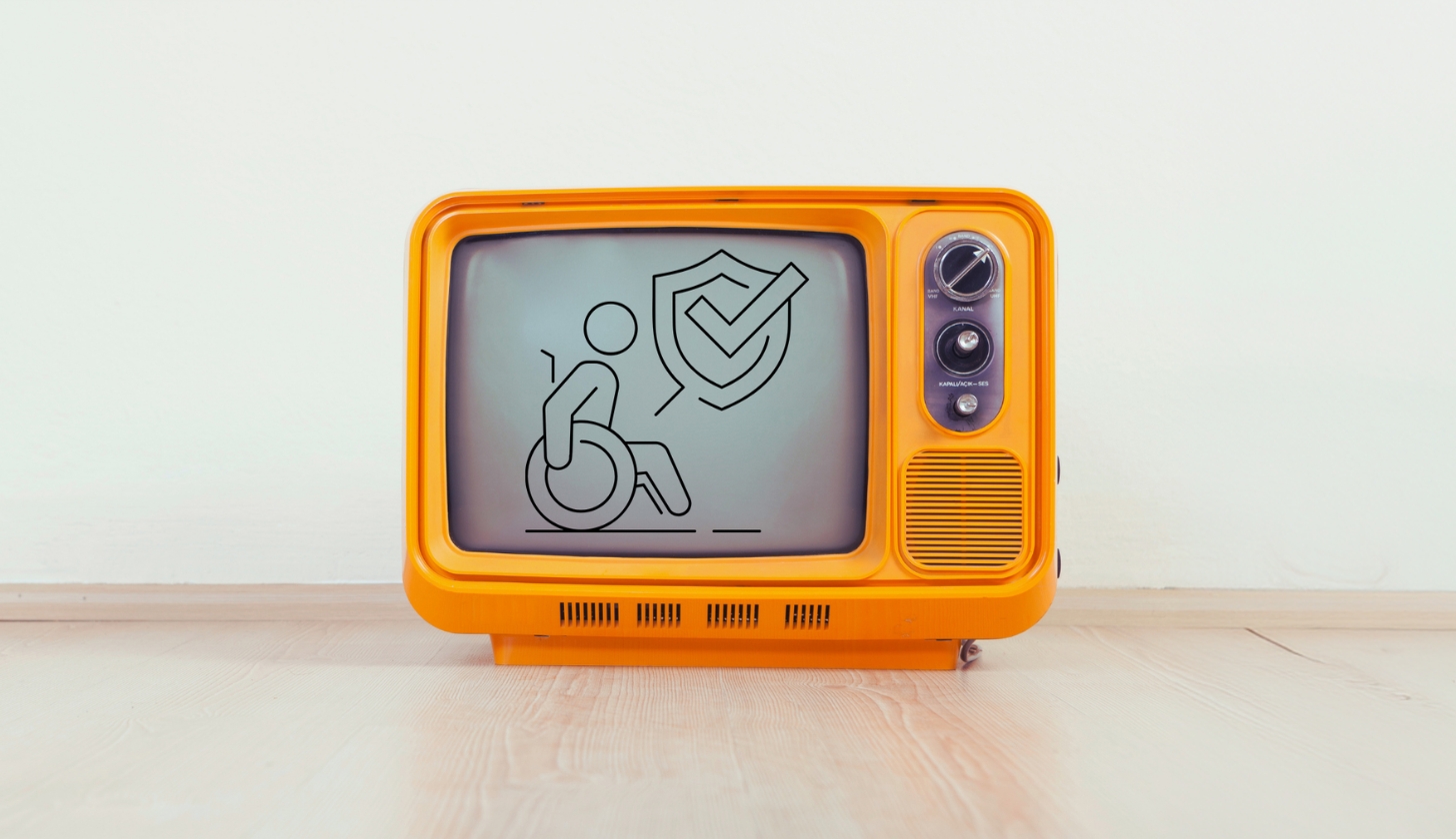 Pomarańczowy telewizor stoi na podłodze. Na jego ekranie jest ikona osoby na wózku i tarczy ze znakiem zatwierdzenia.