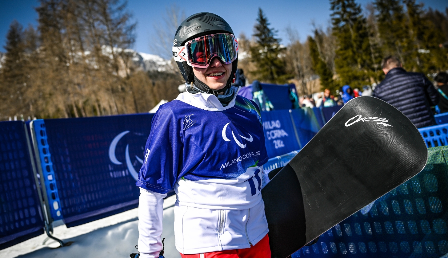 Natalia Siuba-Jarosz w kasku i goglach, ubrana jest w strój sportowy. W ręku trzyma deskę snowboardową. Tło wypełniają drzewa.