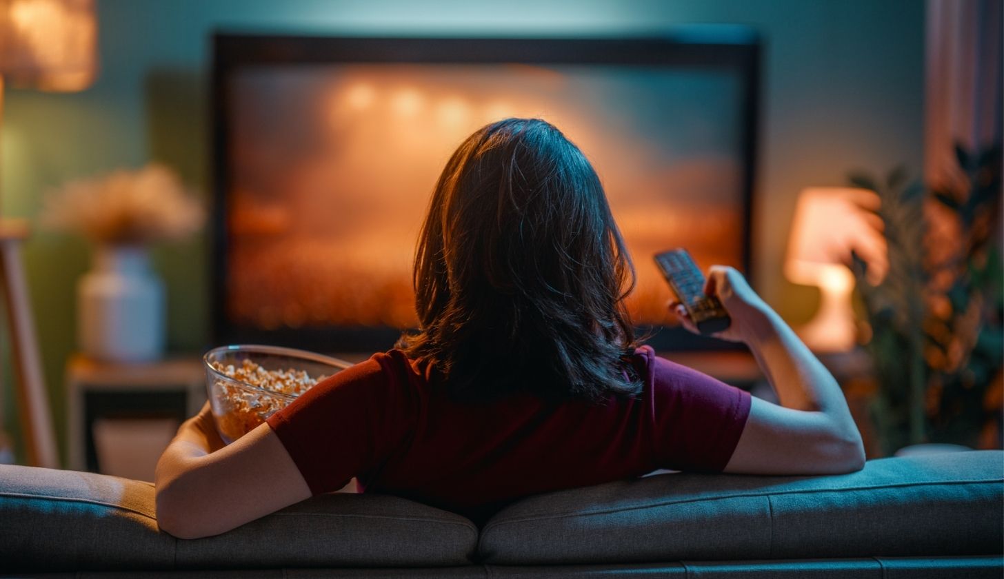 Kobieta siedzi odwrócona tyłem na kanapie i ogląda film. W jednym ręku trzyma popcorn w misce, a w drugim pilot do telewizora.