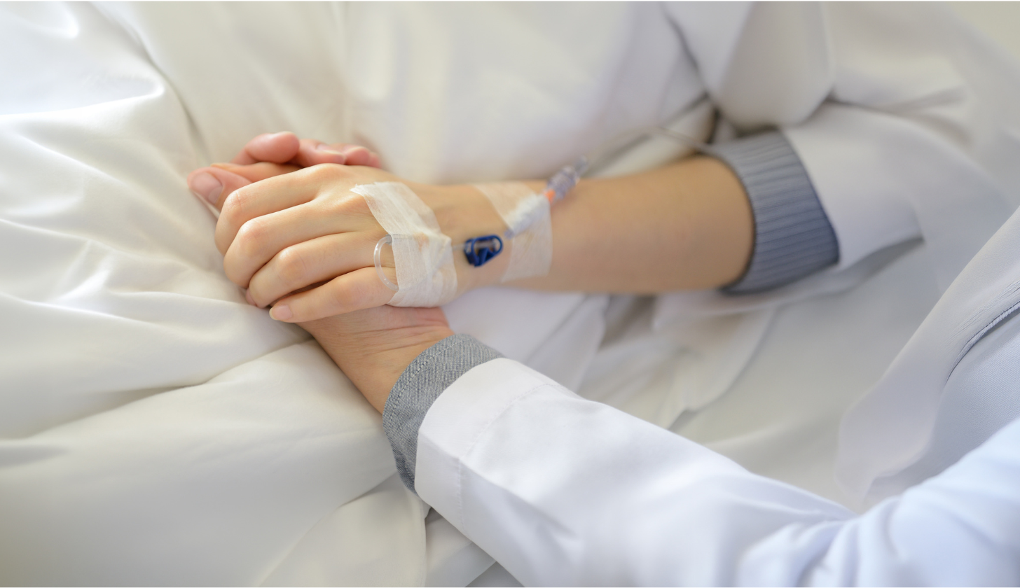 Osoba która leży na łóżku ma podłączoną kroplówkę do dłoni i trzyma za rękę drugą osobę.