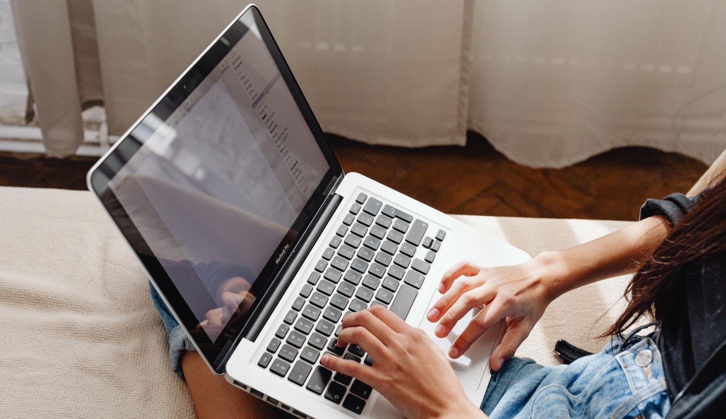 Kobieta w pokoju siedzi na łóżku, trzyma na kolanach laptop. Pisze na klawiaturze.