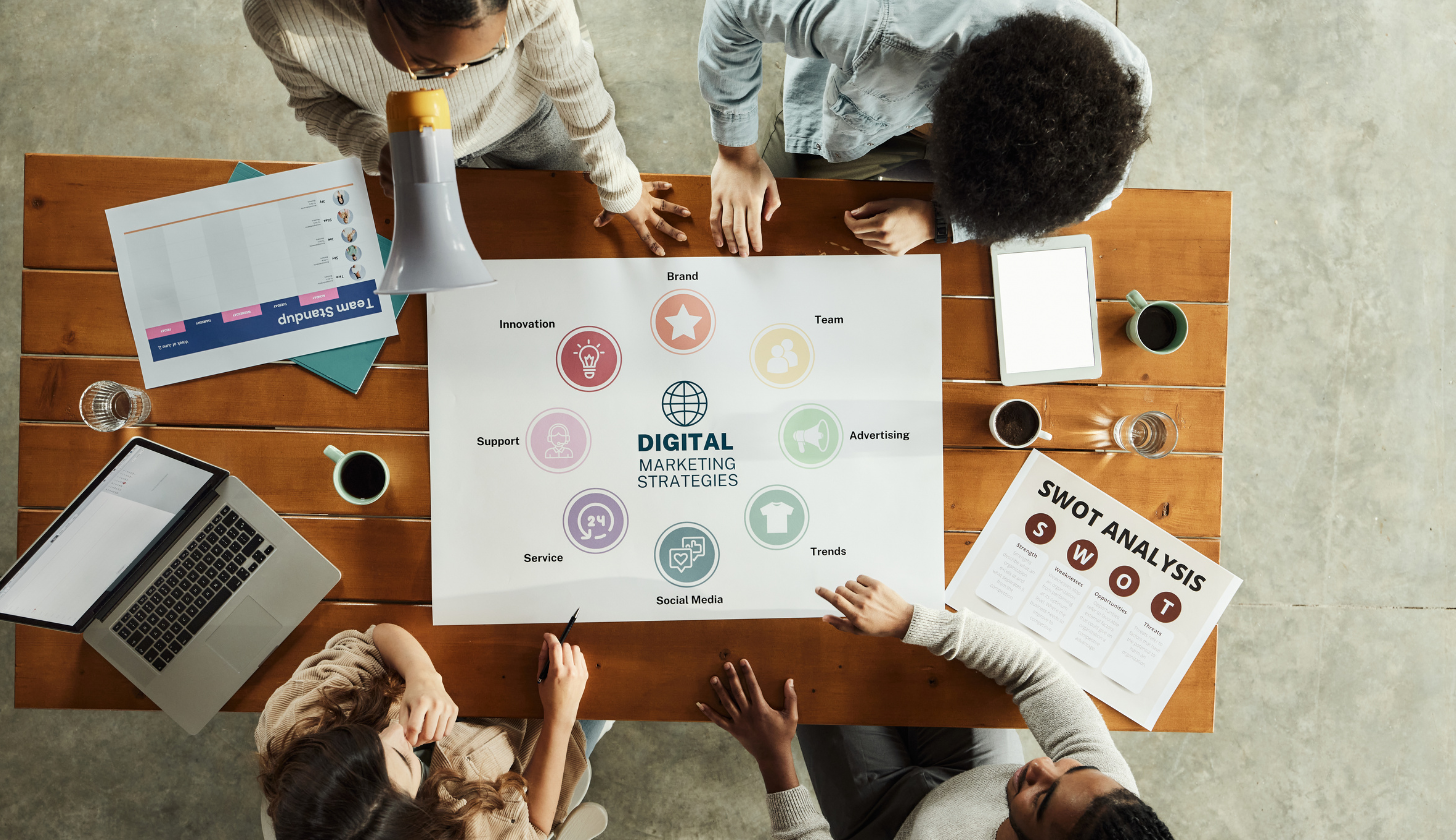 Zdjęcie symbolizuje pracę zespołową nad strategią marketingową. Zdjęcie wykonane z góry przedstawia kilka osób pracujących wspólnie przy dużym drewnianym stole. W centrum leży plansza