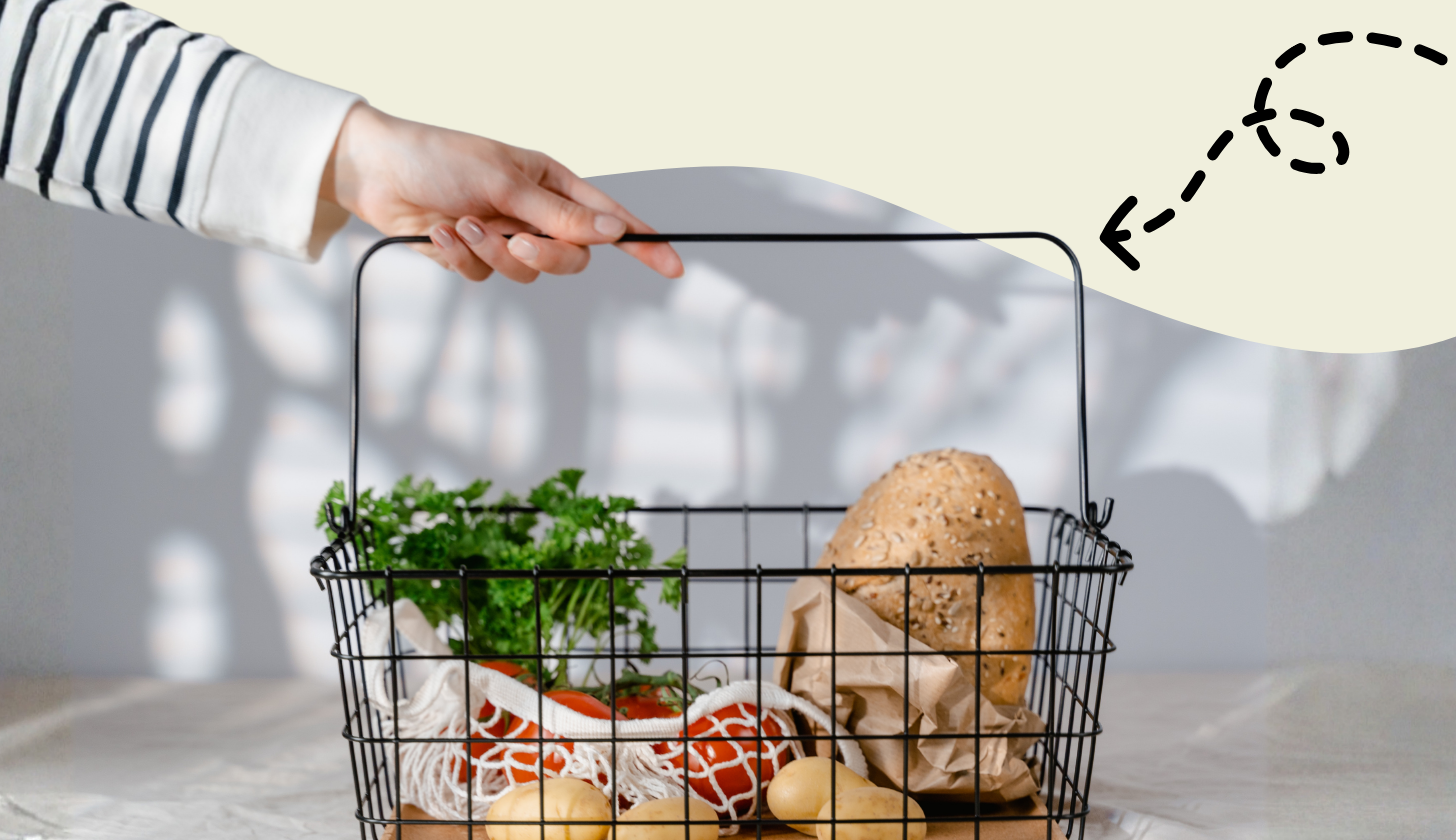 Dłoń chwyta za uchwyt koszyka zakupowego w którym są produkty zielona pietruszka, pomidory, ziemniaki i chleb.