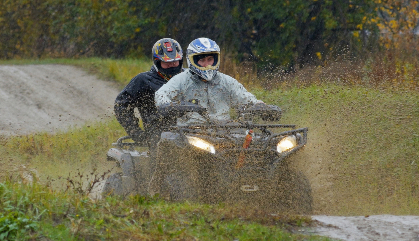 Na drodze nieutwardzonej, dwie osoby poruszają się quadem. Ubrani są w kaski i kurtki. W czasie jazdy błoto pryska pod kołami.